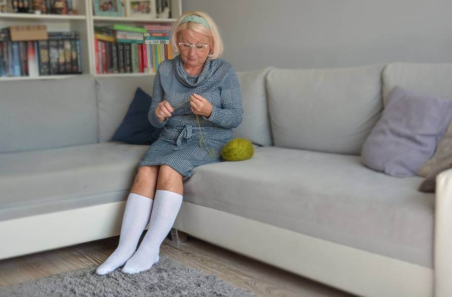 An elderly woman on a couch, knitting, with Rainbow high-knee socks.